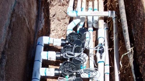 A close-up view of PVC pipes and drip emitters used in an irrigation system installed in a trench in the dirt.