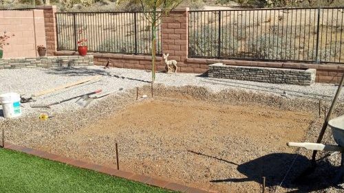 An aerial or elevated view of a backyard under construction, showing a large area prepped with dirt and gravel, surrounded by a fence and a strip of new synthetic grass.