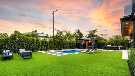 A modern backyard featuring a swimming pool, covered patio, and a perfectly green, lush area of synthetic grass lit up at sunset.